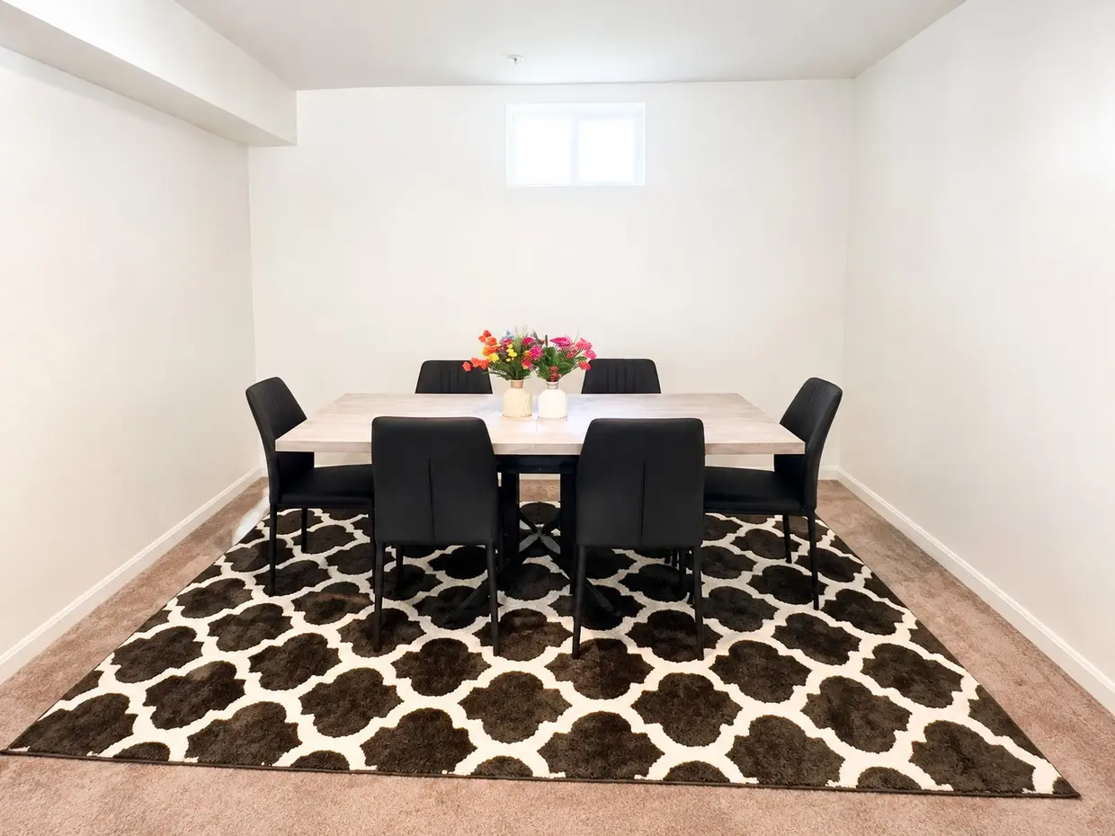 Dining room with a table for six and vases of flowers on a patterned rug, beneath a small window.